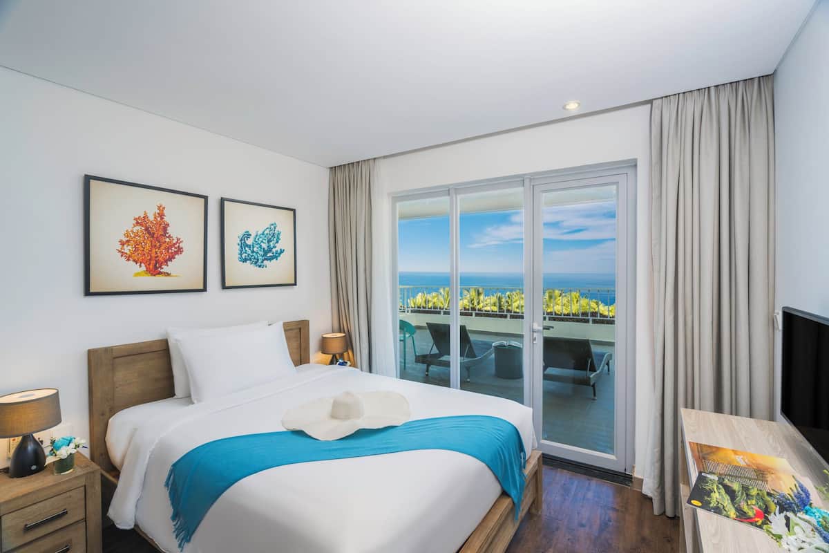 A hotel room with a bed, a TV, a desk, and a balcony with a view of the ocean.