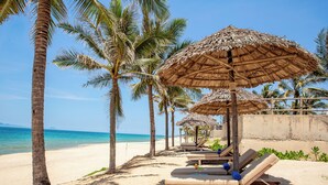 A beach with palm trees, sun loungers, and thatched umbrellas. - The Pearl Hội An (Da Nang)