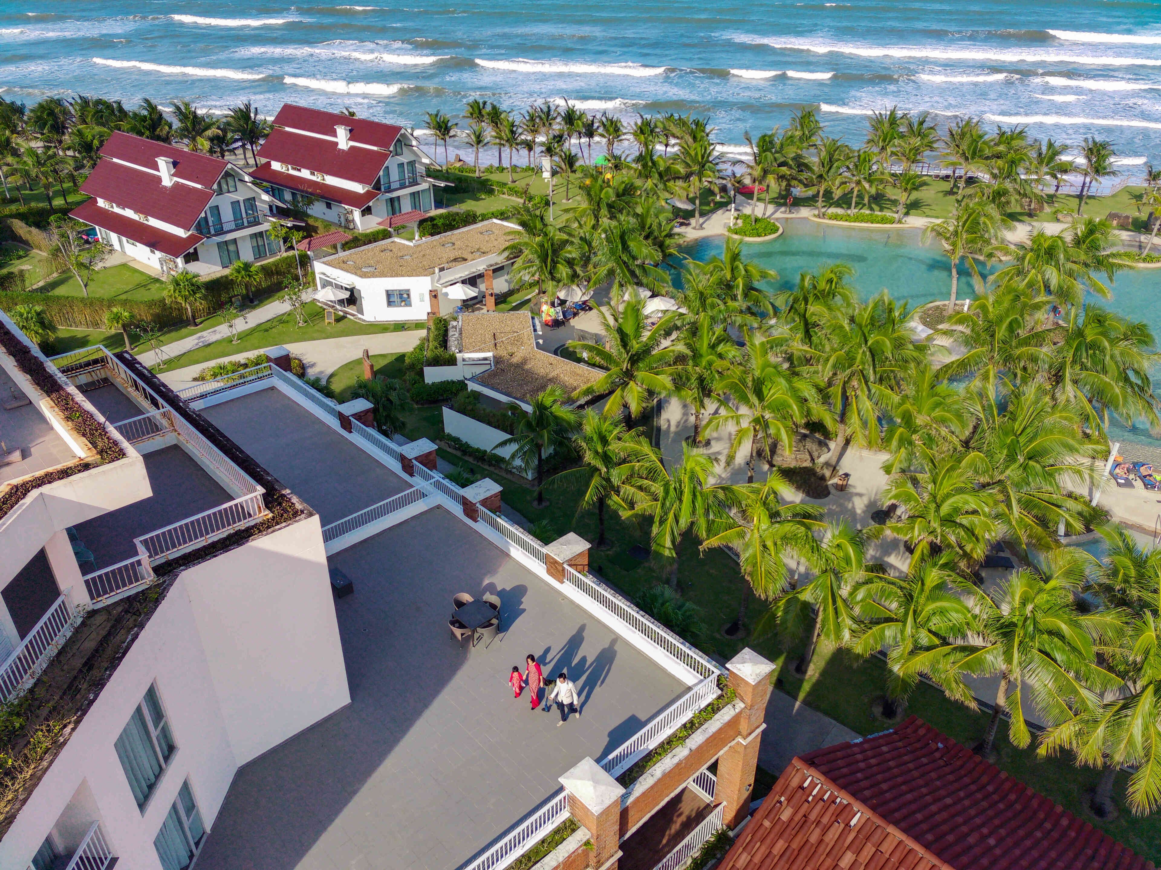 An aerial view of a resort with multiple buildings, a pool area surrounded by palm trees, and a view of the ocean.