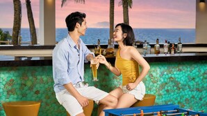 A couple sitting at a bar with a view of the ocean, a foosball table, and a bar stocked with bottles. - The Pearl Hội An (Da Nang)
