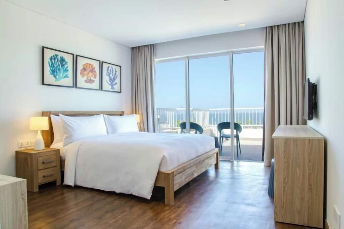 A hotel room with a bed, bedside table, lamp, artwork, and a balcony with chairs.