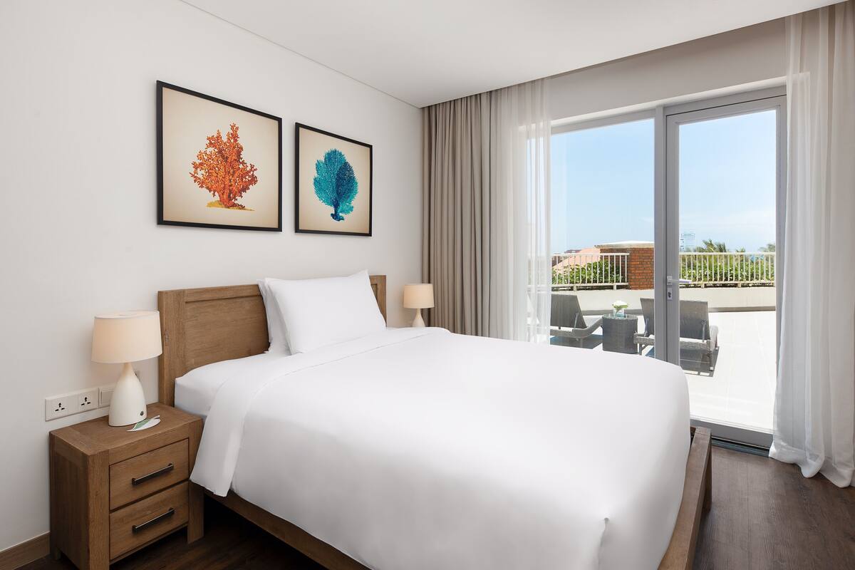 A hotel room with a large bed, two bedside tables with lamps, a view of the outdoor area through a sliding glass door, and two framed artworks on the wall.