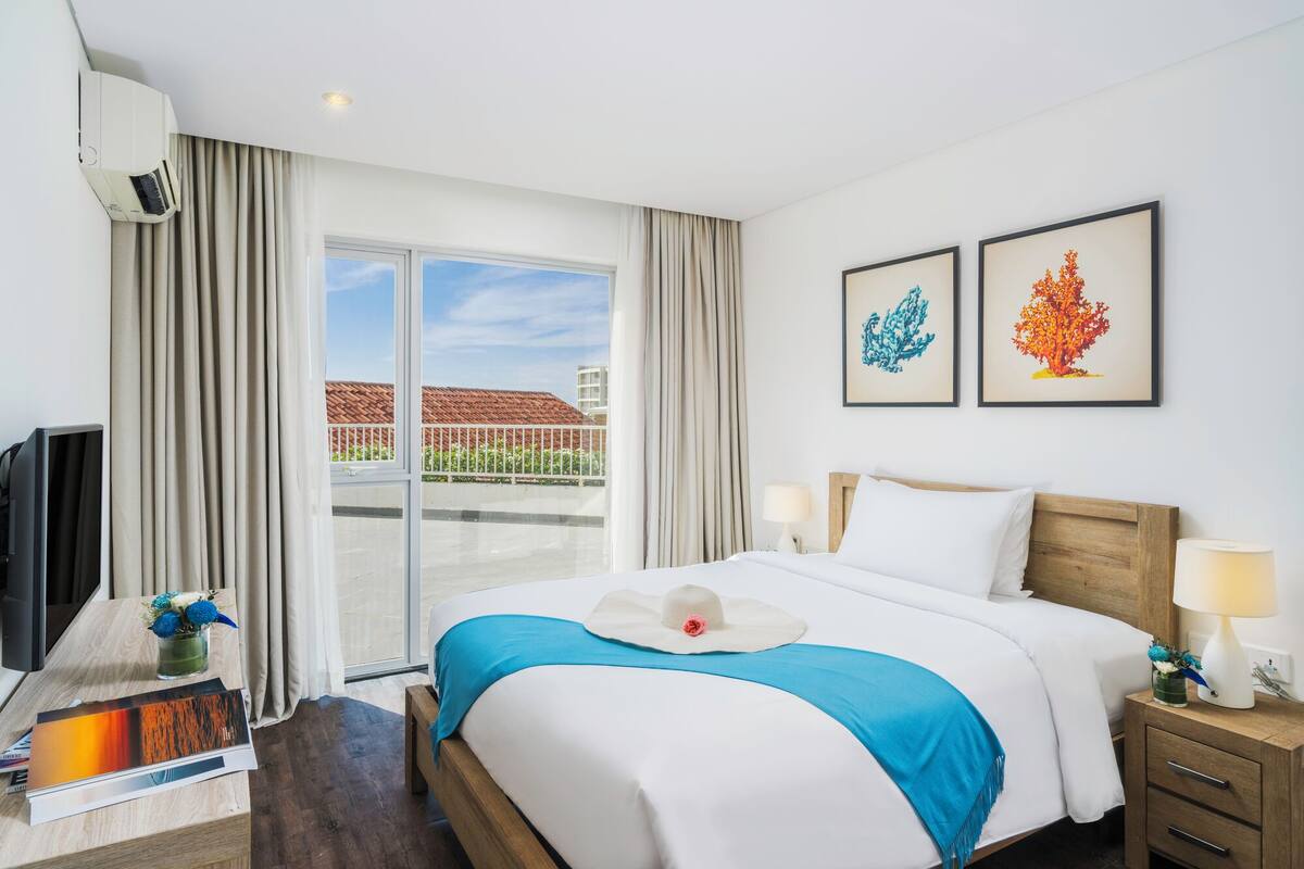 A hotel room with a bed, bedside tables, a TV, and a balcony with a view of buildings.