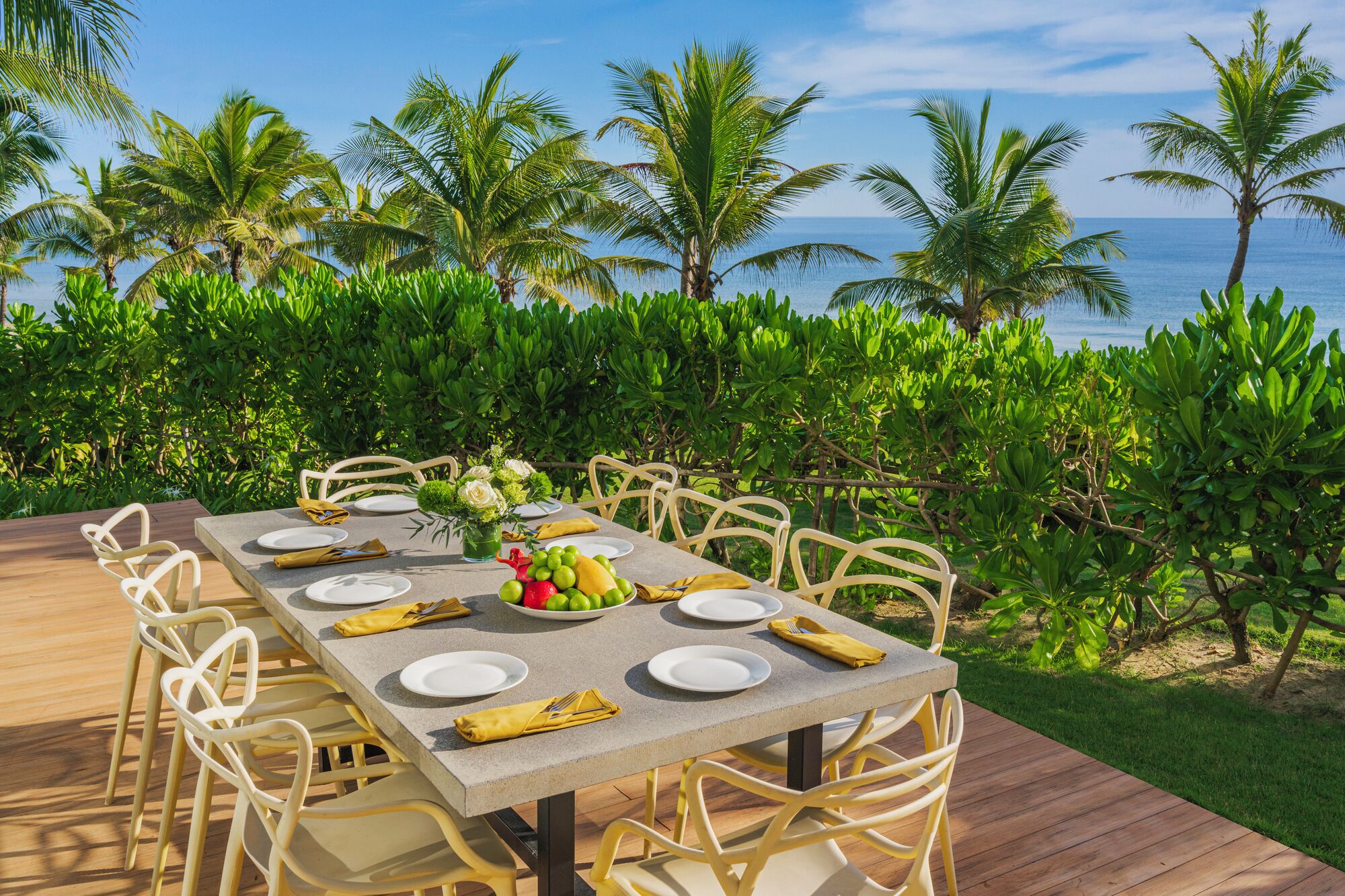 An outdoor dining area with a view of the ocean, surrounded by lush greenery and palm trees.
