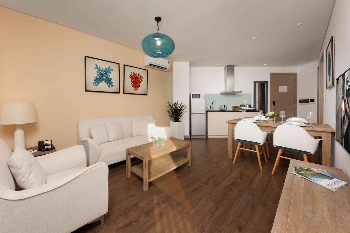 A modern living room with a sofa, coffee table, and dining area. There is a kitchen in the background with a microwave and stove.