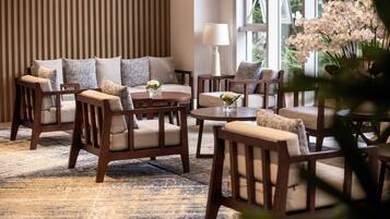 A modern lounge area with sofas and armchairs arranged around a central coffee table, a patterned rug, and a large window providing a view of greenery.