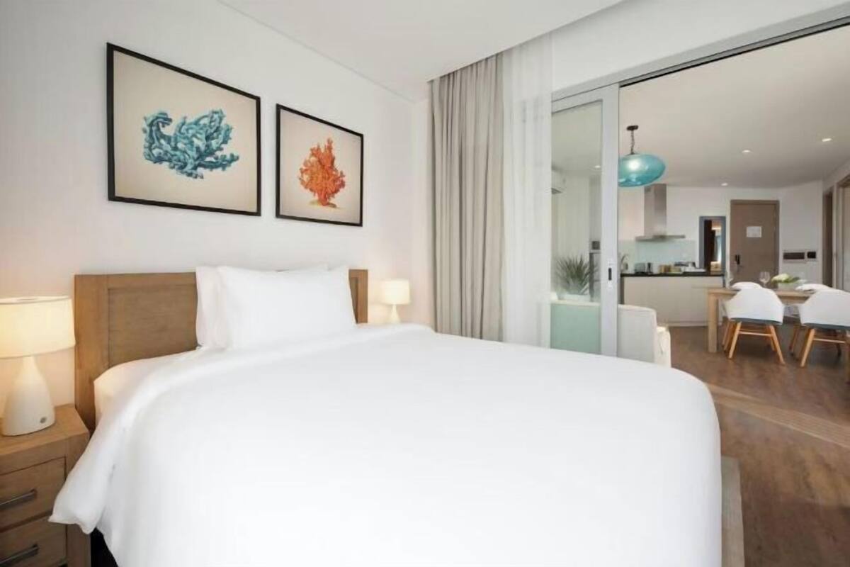 A hotel room with a bed, two framed artworks, a nightstand, a lamp, a sliding glass door leading to a dining area with chairs and a table.