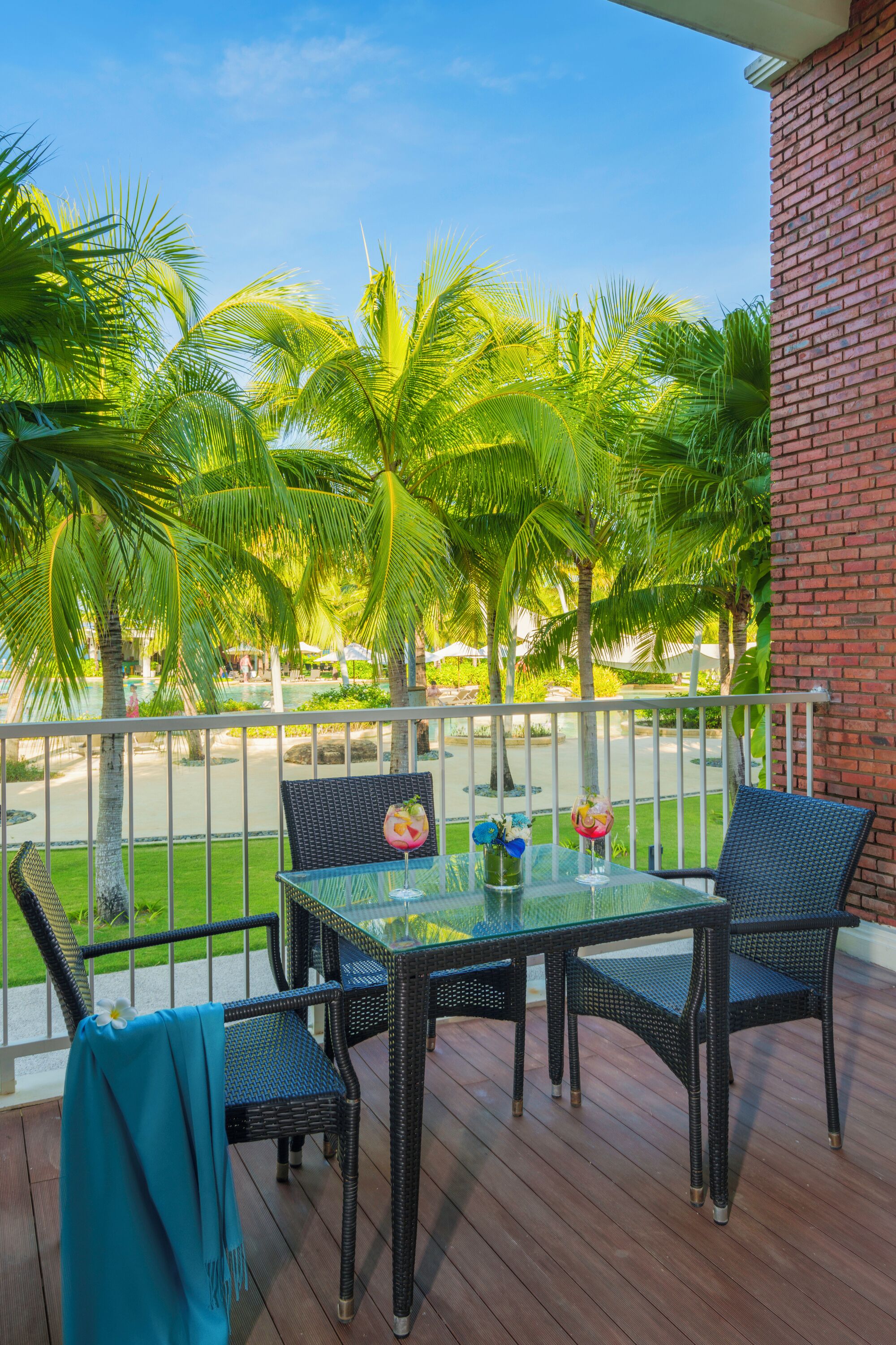 A balcony with a glass table and wicker chairs, overlooking a pool and palm trees.