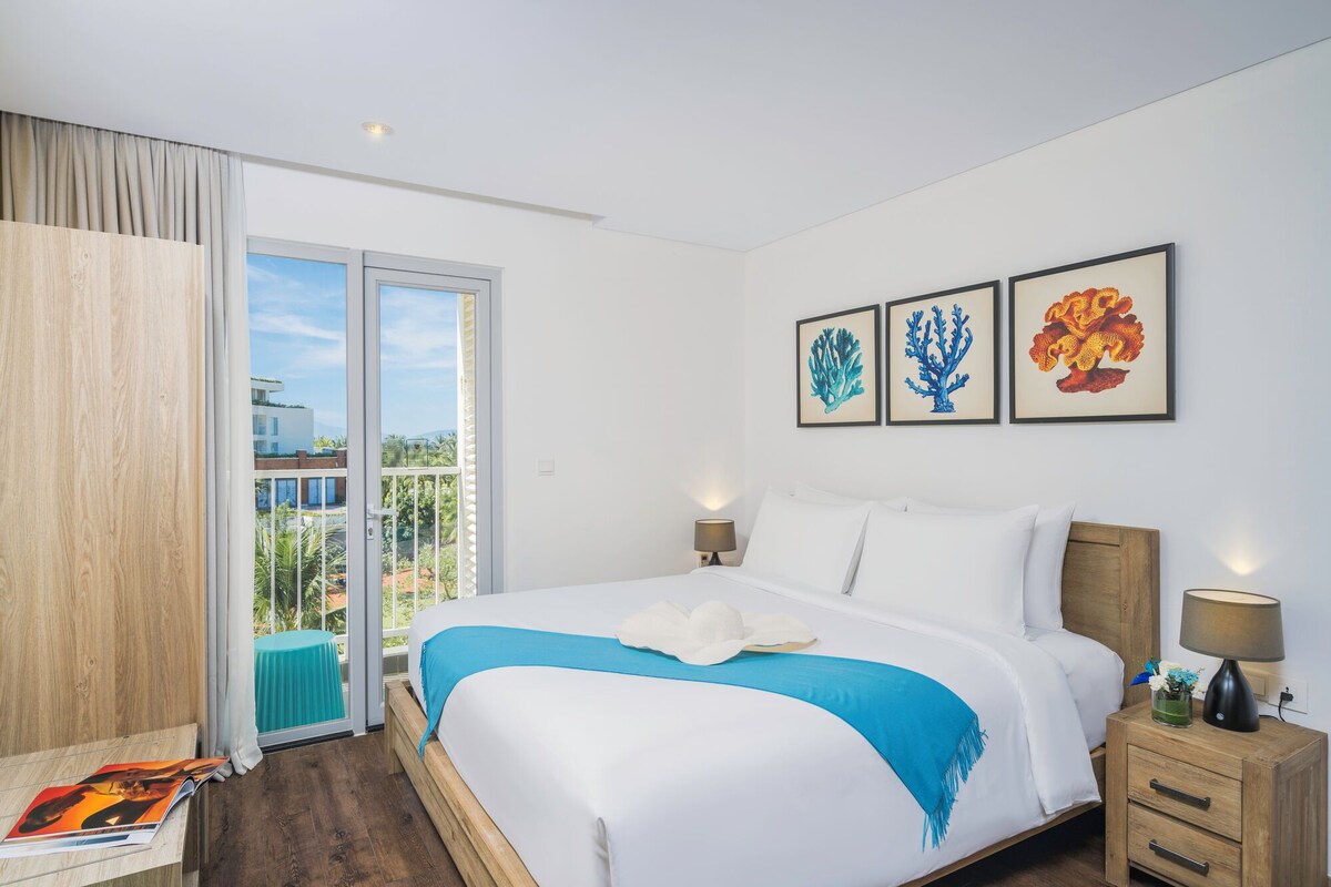 A hotel room with a large bed, a nightstand, a window with curtains, and three framed artworks on the wall.