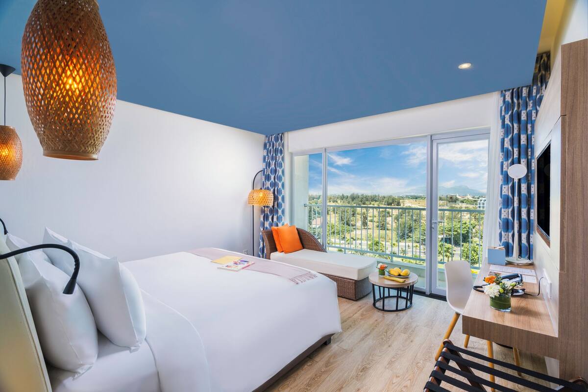 A modern hotel room with a large bed, a desk, and a balcony with a city view.