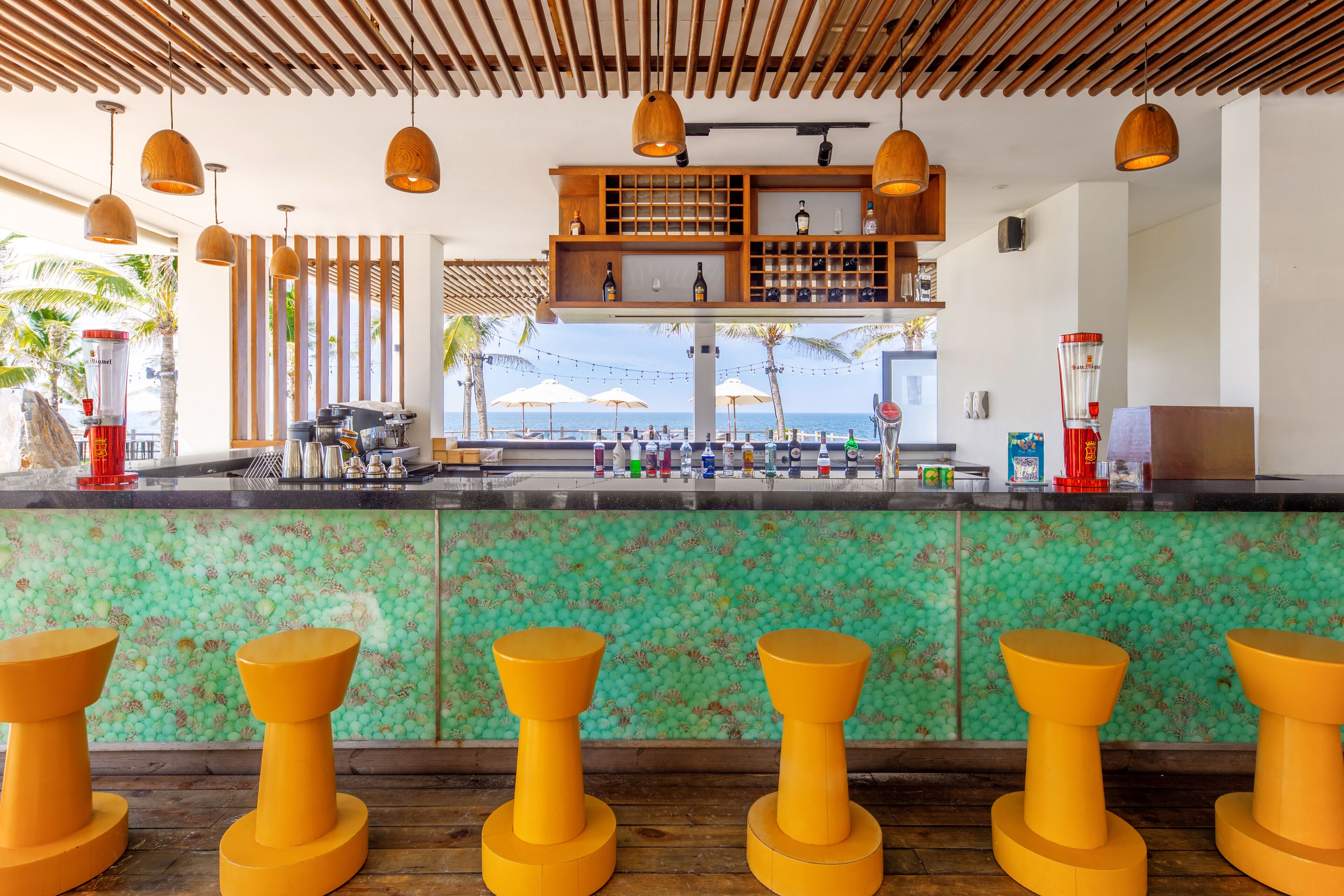 A bar area with a green mosaic backsplash, wooden slatted ceiling, and yellow stools.