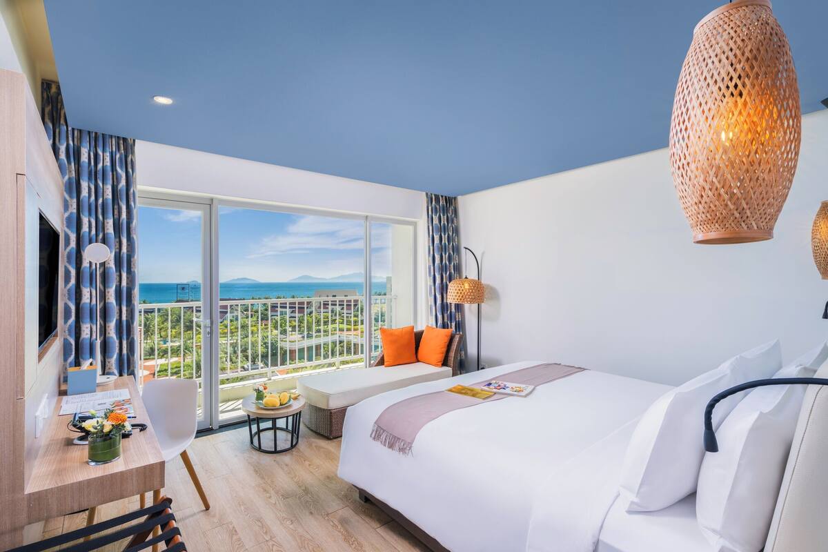 A hotel room with a large bed, a desk with a chair, a small table with flowers, and a view of the ocean through a large window.