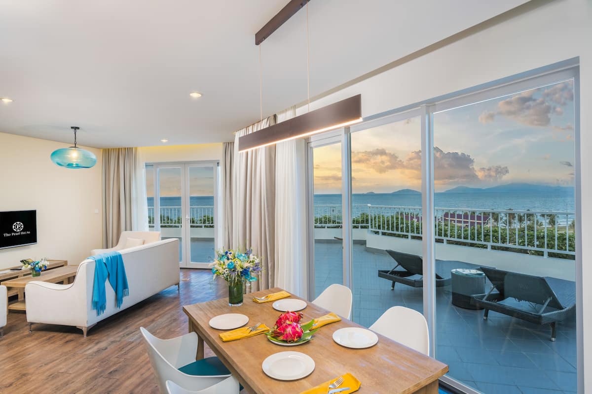 A modern living room with a dining area, large windows with a view of the sea, and a flat-screen TV mounted on the wall.