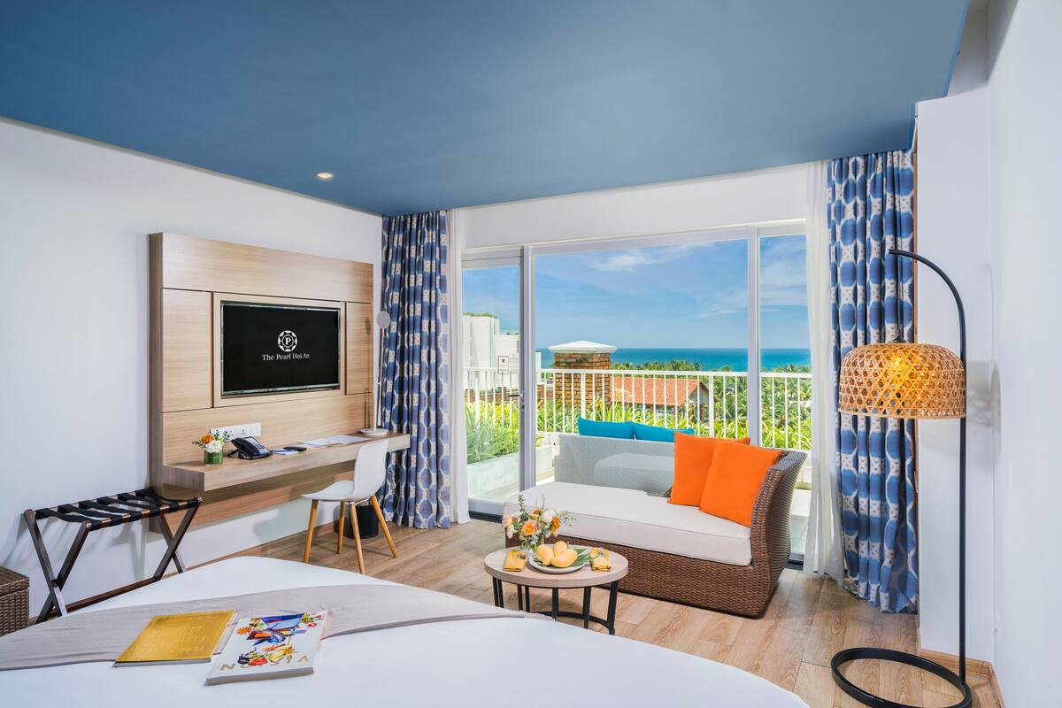 A hotel room with a balcony, a TV, a desk, a sofa, and a dining table.