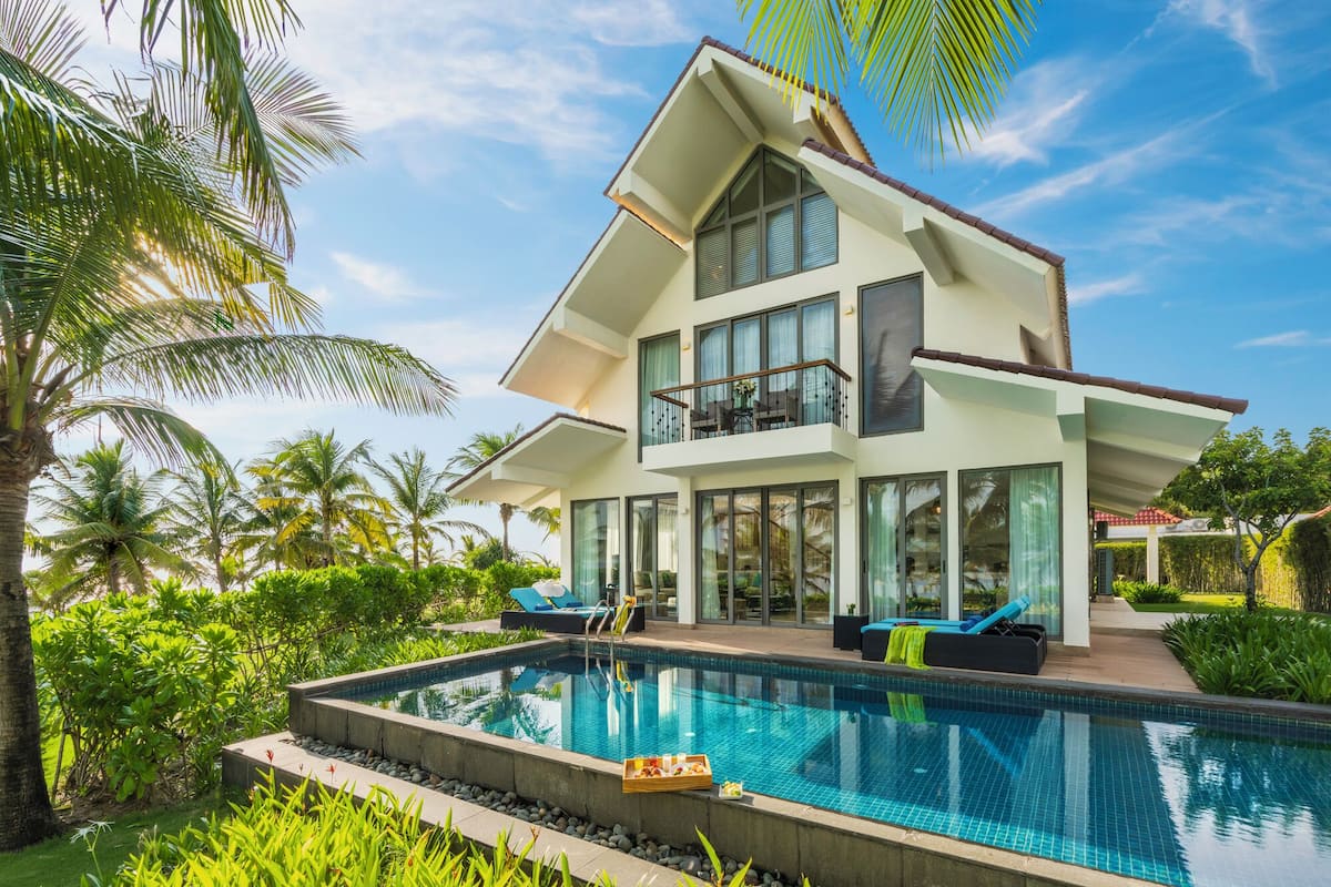 A modern two-story house with a large swimming pool, surrounded by lush greenery and palm trees.