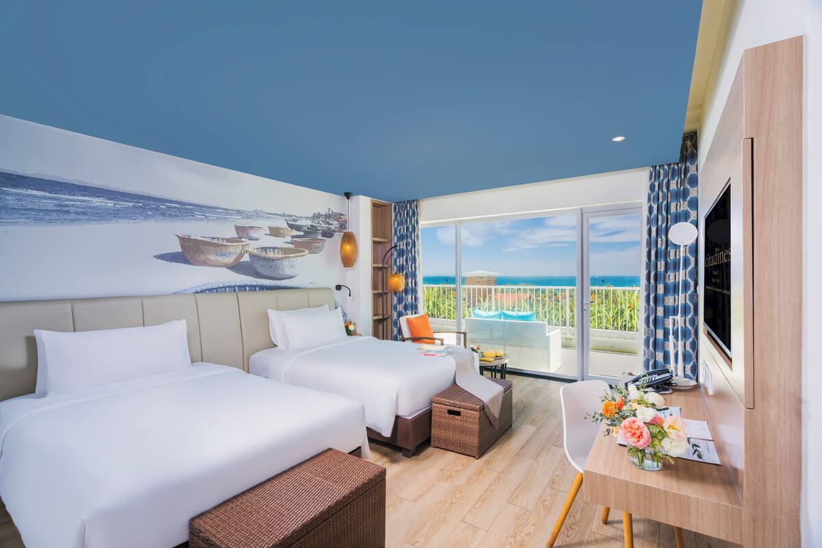 A hotel room with two beds, a desk with a vase of flowers, a TV, and a balcony with a view of the sea.