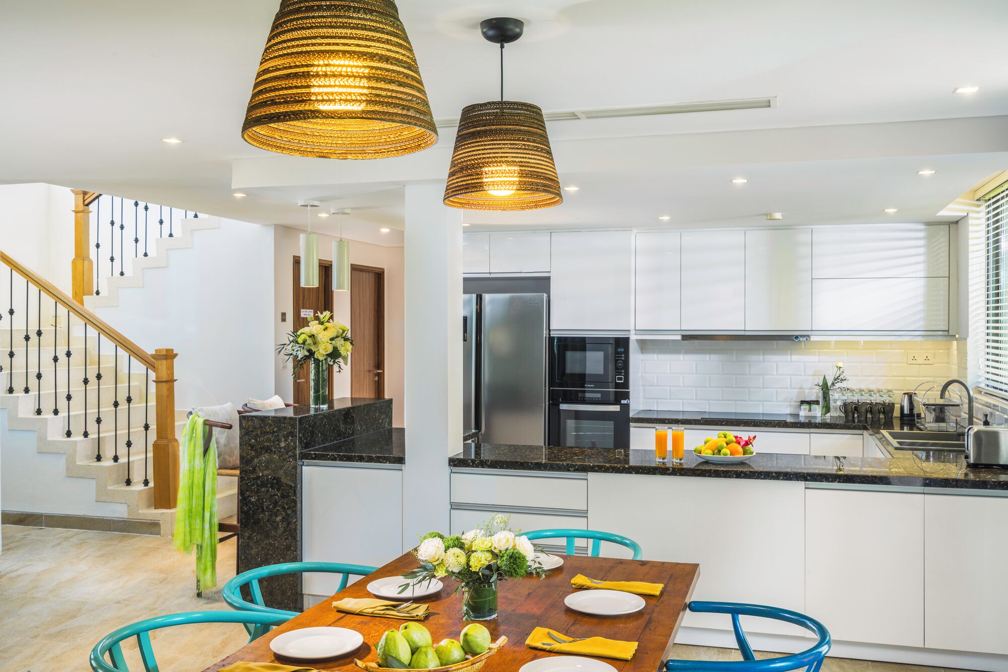 A modern kitchen with white cabinets, a stainless steel refrigerator, and a black countertop. There is a dining table with chairs and a vase of flowers.