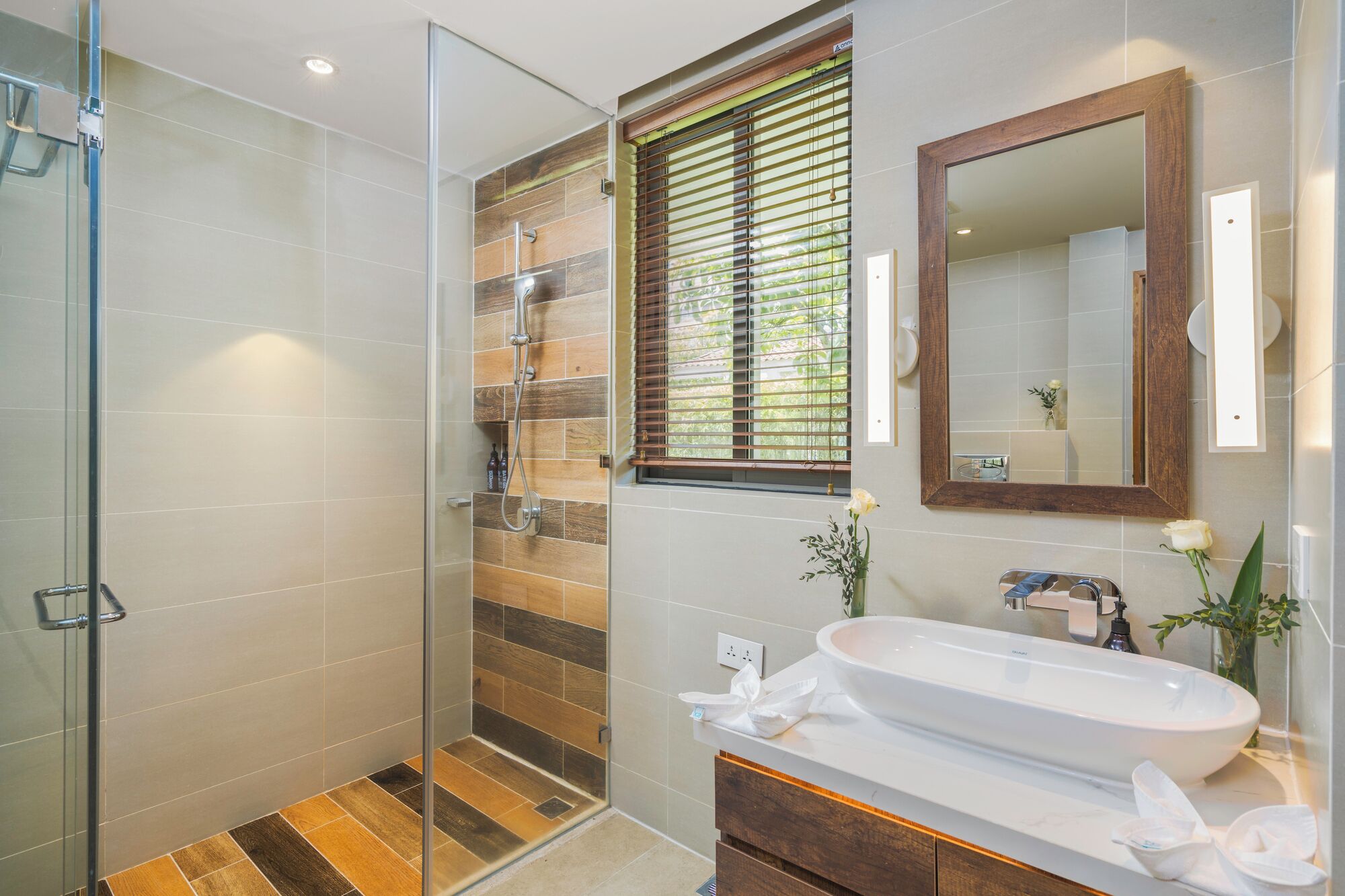 A modern bathroom with a glass shower enclosure, a wooden-tiled shower area, a large mirror, and a white sink with a vase of flowers.