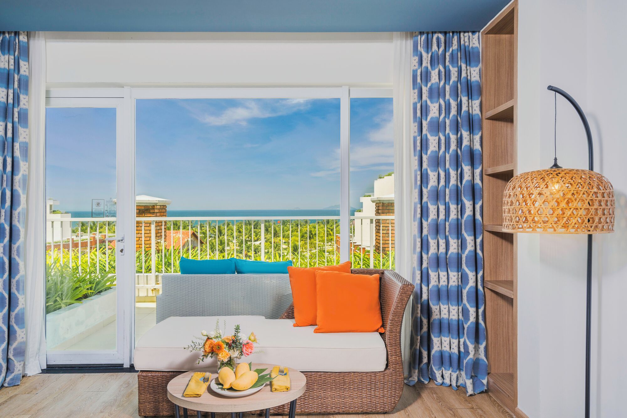 Een balkon met uitzicht op zee, een rieten zithoek met kussens, een bijzettafeltje met fruit en bloemen, en een hanglamp.