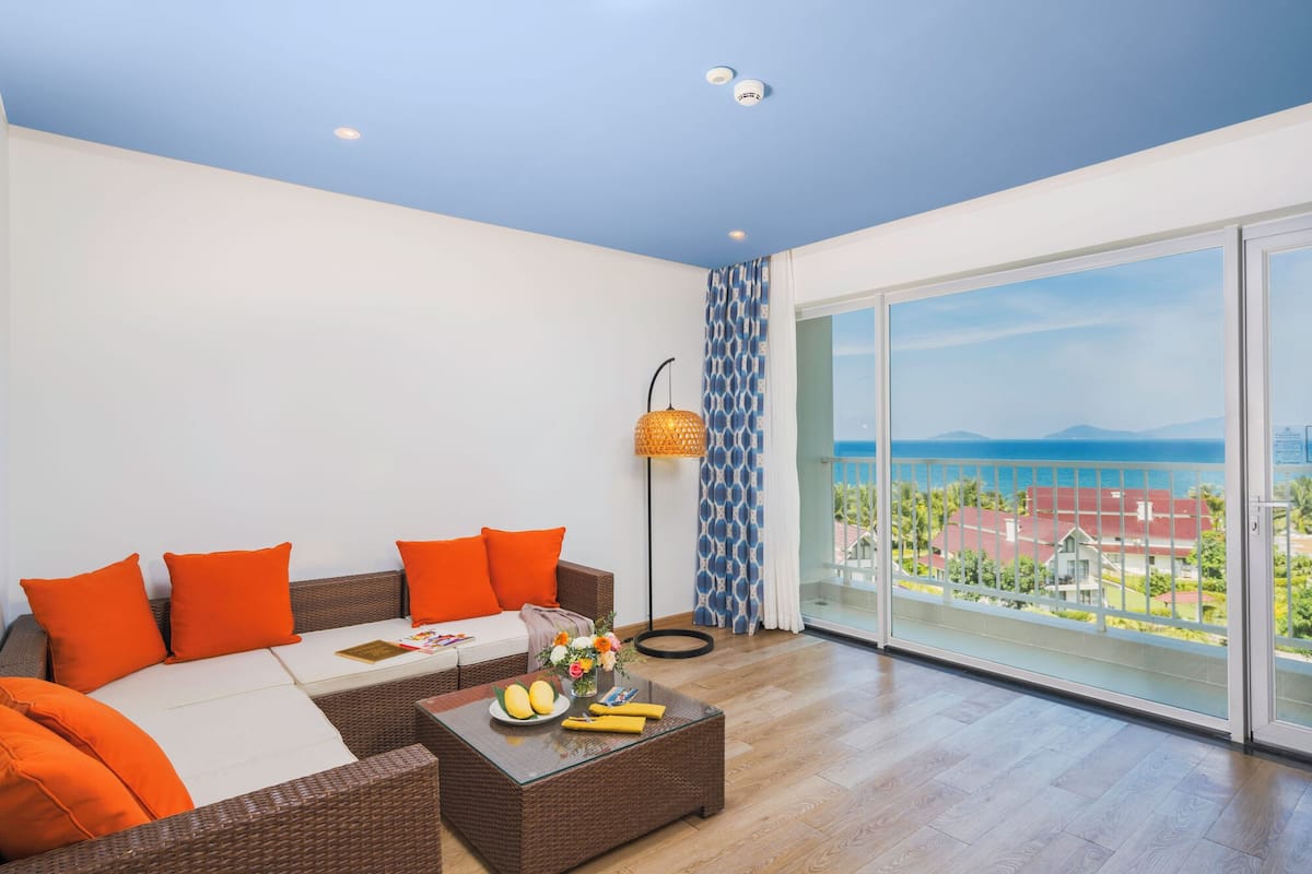 A modern living room with a large glass door leading to a balcony with a view of the sea and buildings.