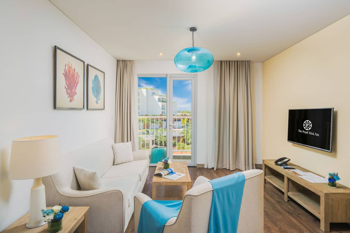 A hotel room with a sofa, armchairs, a coffee table, a TV, and a balcony with curtains.