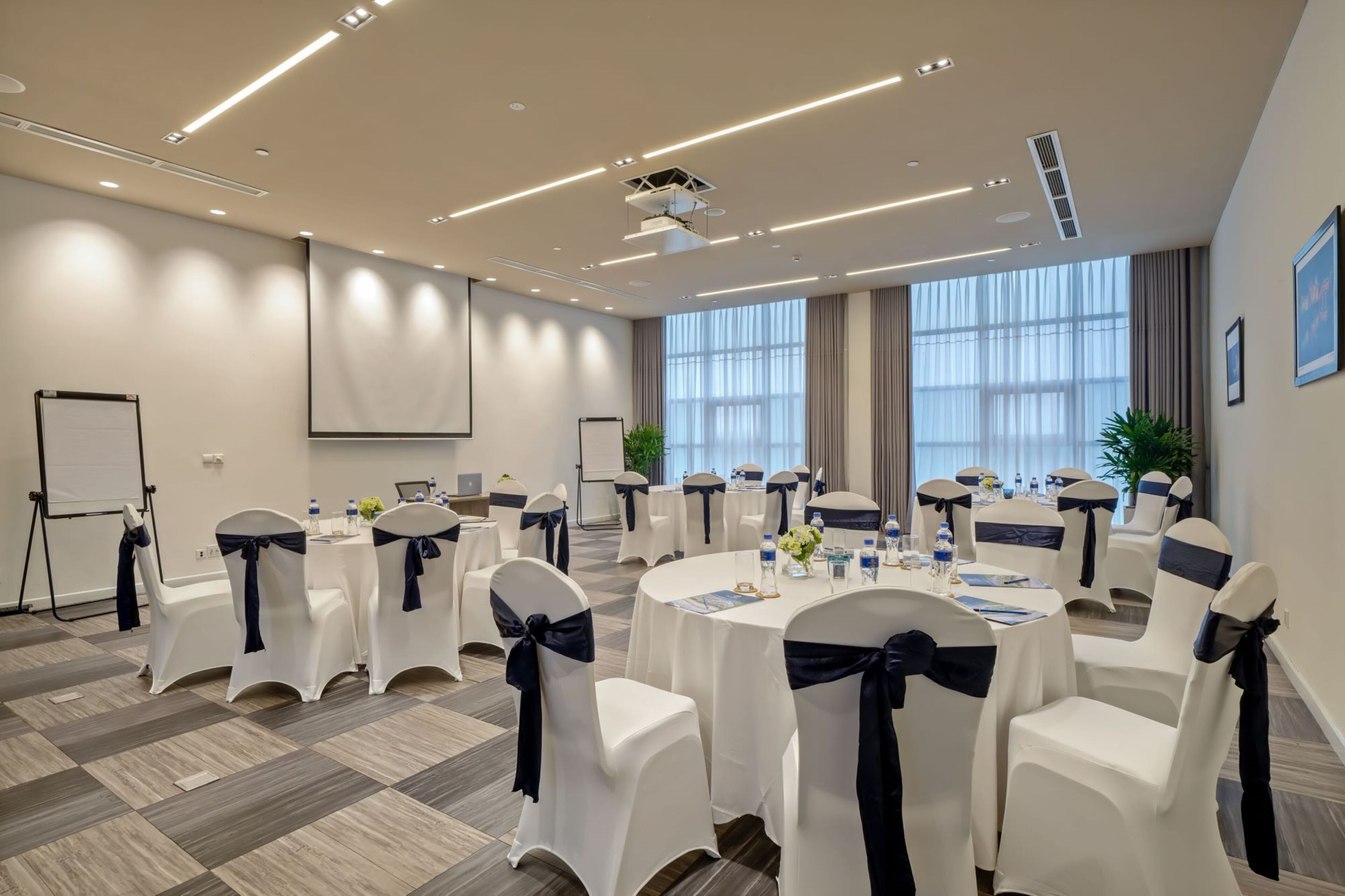 Une salle de conférence équipée de tables rondes, de chaises blanches avec des rubans foncés, d’un écran de projection et d’un tableau blanc.