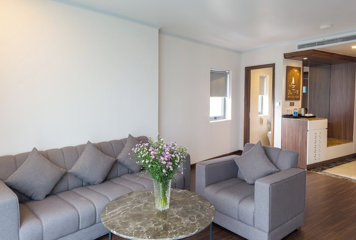 Uma sala moderna com um sofá modular cinza, uma mesa de centro de mármore redonda e um vaso com flores. Há uma janela pequena e uma porta que dá para uma área de cozinha compacta.