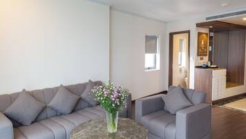 Uma sala moderna com um sofá modular cinza, uma mesa de centro de mármore redonda e um vaso com flores. Há uma janela pequena e uma porta que dá para uma área de cozinha compacta.