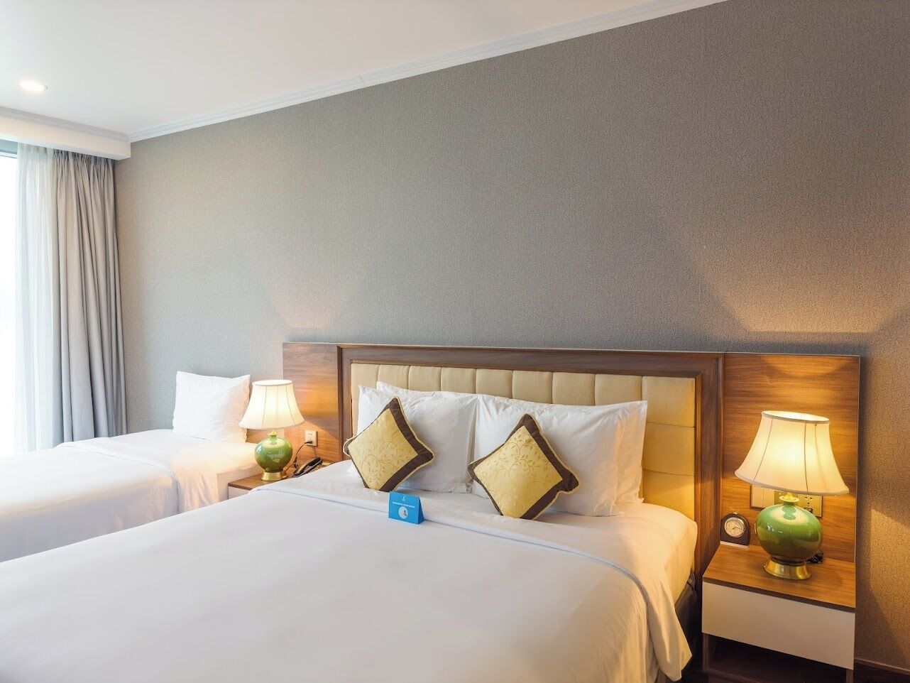 A hotel room with two beds, a wooden headboard, beige walls, and green lamps.