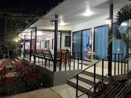 Un edificio con una zona exterior techada, escalones que conducen a un balcón y un área de comedor con mesas y sillas.