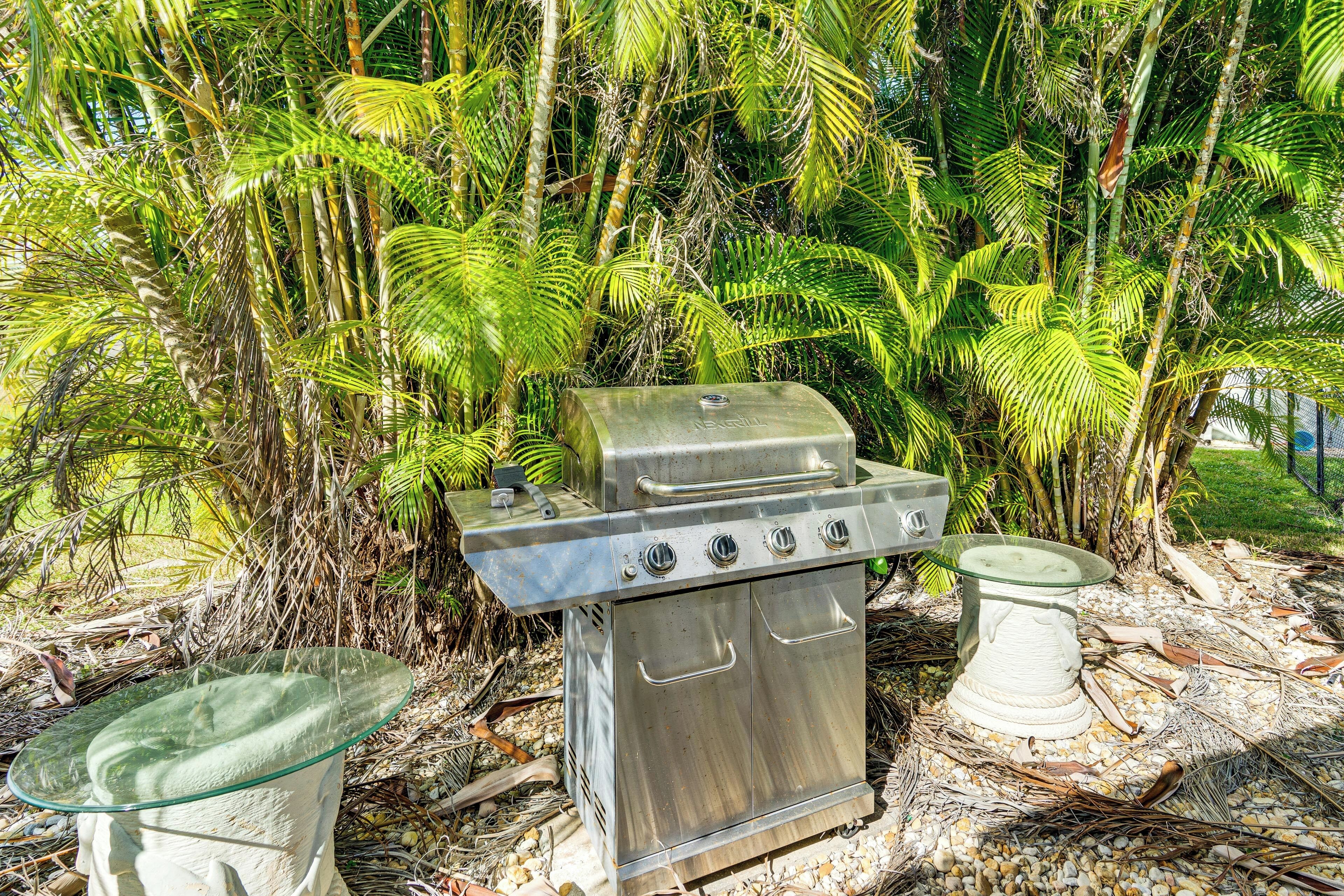 A stainless steel barbecue grill with a glass top and two side tables, set in a garden with tropical plants.