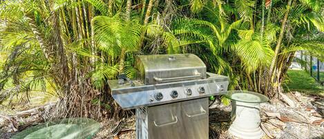 A stainless steel barbecue grill with a glass top and two side tables, set in a garden with tropical plants.
