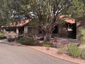 A rustic house with a large tree in front, a stone wall, and a red-tiled roof.