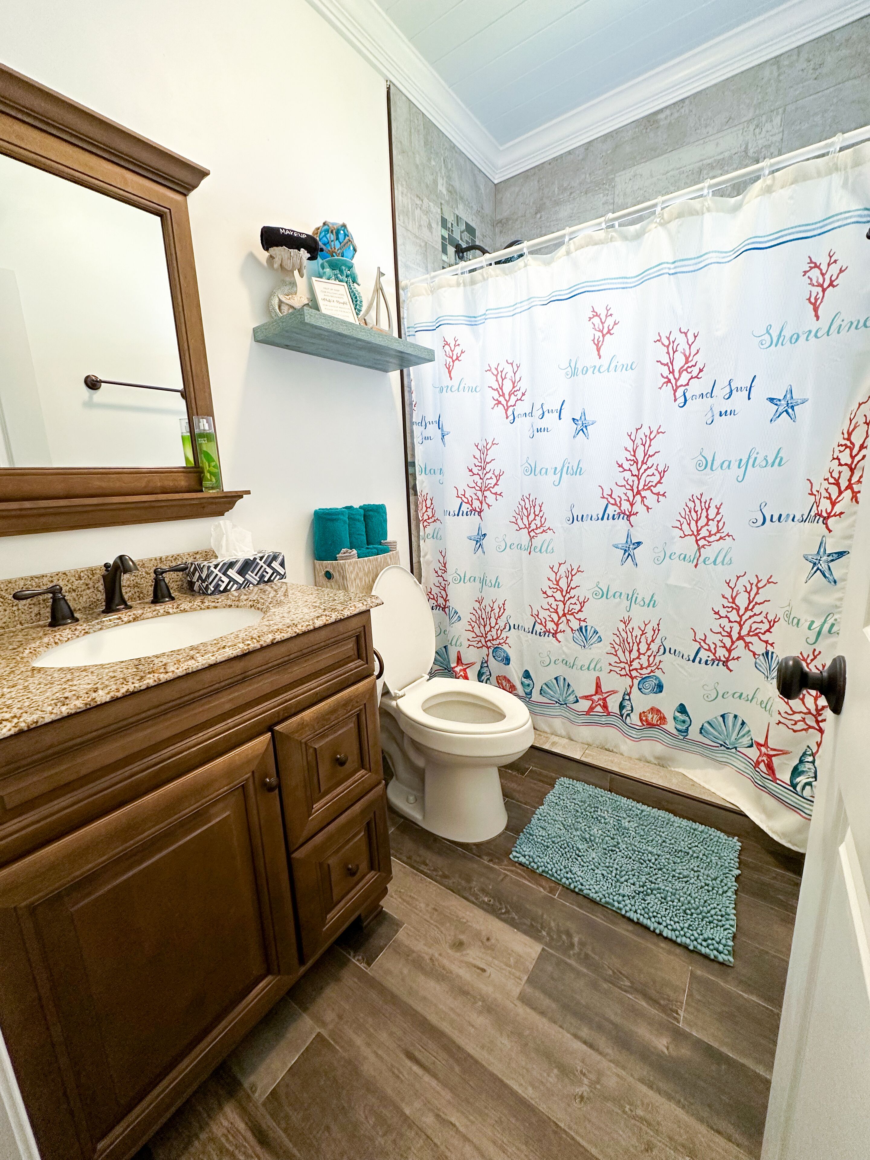 A bathroom with a wooden vanity, a white toilet, a shower curtain with sea-themed designs, and a blue bath mat.