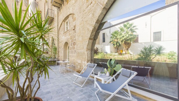 A courtyard with stone walls, two lounge chairs, a potted plant, and a table with a chair. - Luxury Gattopardo Loft - LAGO Design by Wonderful Italy (Palermo)