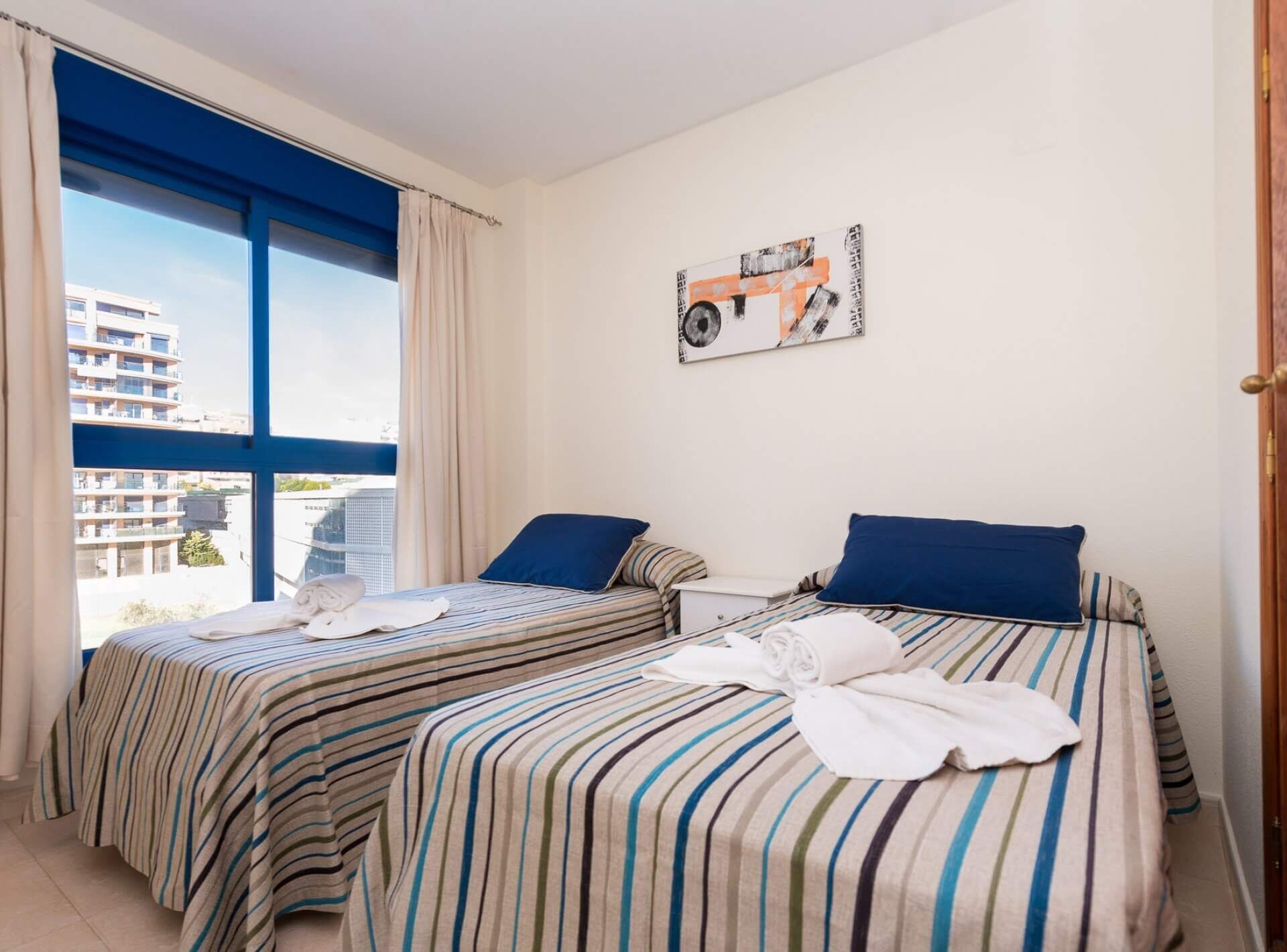 A hotel room with two beds, striped bedding, a window with curtains, and a city view.