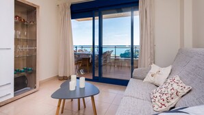 Un soggiorno moderno con divano, un tavolino con candele, una zona pranzo con sedie e un tavolo e vista sul mare attraverso una porta vetrata.