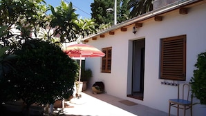 Uma pequena área de pátio com um edifício branco, um guarda-sol riscado em vermelho e branco, e uma cadeira de madeira.