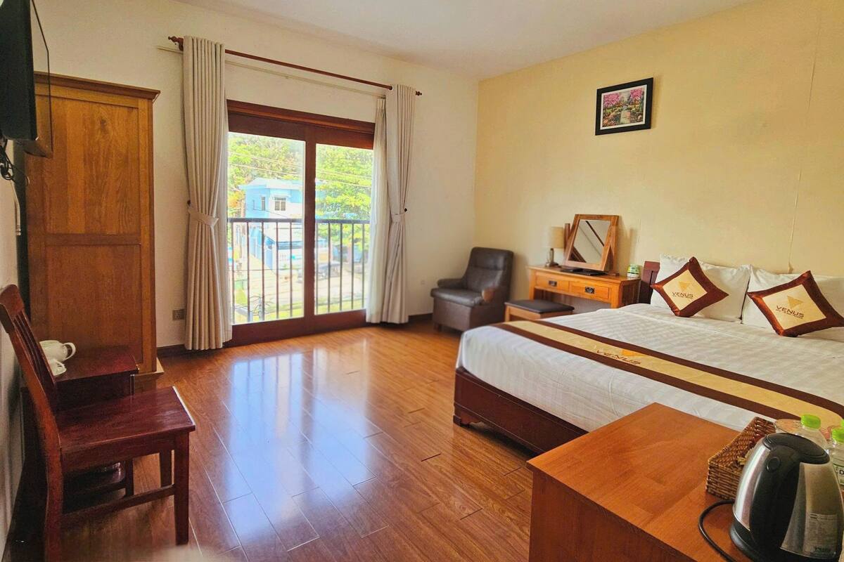 A hotel room with a bed, a desk, a chair, a wooden wardrobe, and a view of the outside.