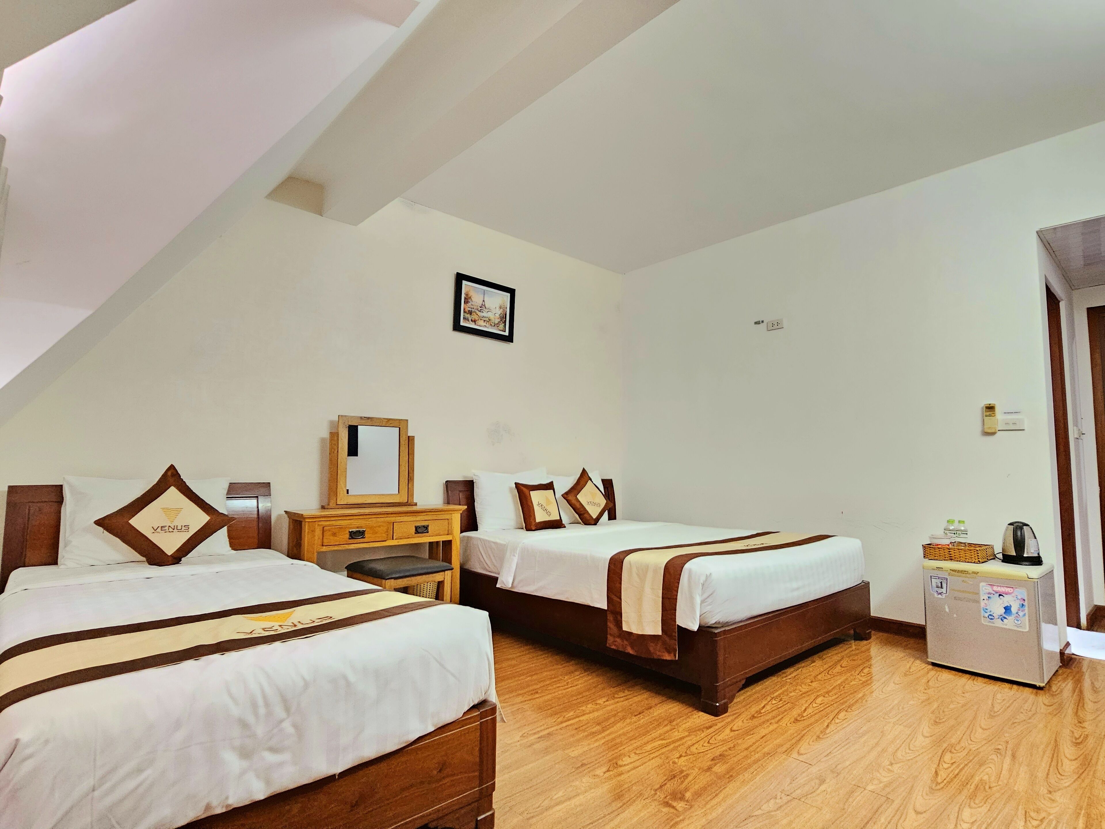 A hotel room with two beds, a wooden dresser, a small table, a refrigerator, and a picture on the wall.