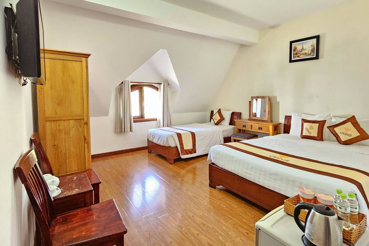 A hotel room with two single beds, a wooden desk, a television, a window with curtains, a wooden wardrobe, and a framed picture on the wall.