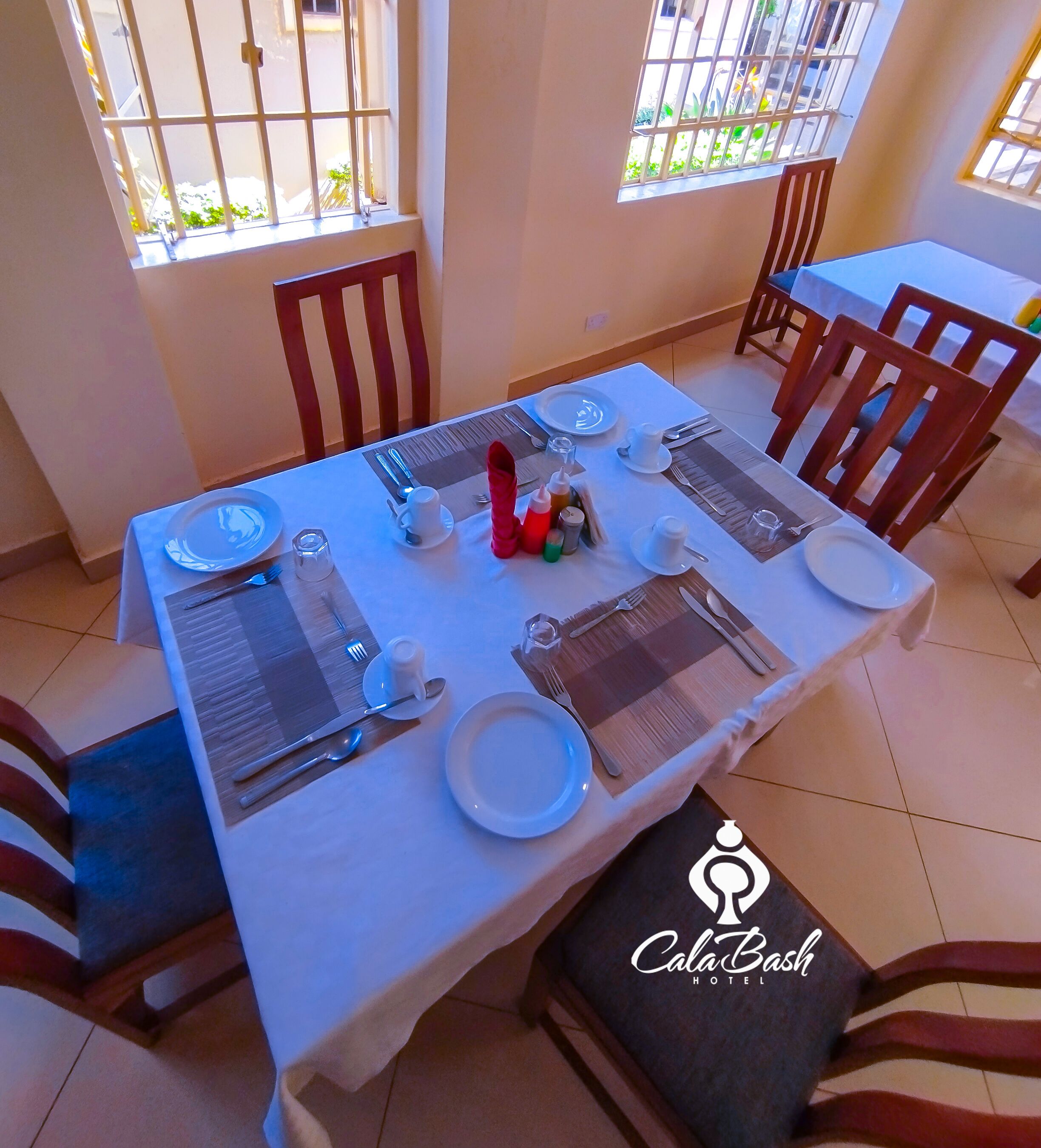 A dining area with a table set for four, including plates, cutlery, and glasses. There are chairs around the table, and a window with bars is visible.