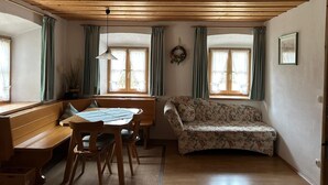 A cozy room with a wooden ceiling, a dining table with chairs, a floral sofa, and two windows with curtains. - "Zellerhorn" Ferienwohnung I°, 39 qm, extra Schlafzimmer° (Aschau im Chiemgau)