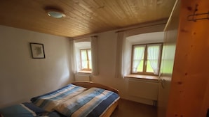 A bedroom with a wooden ceiling, two windows with curtains, a bed with striped bedding, and a radiator. - "Zellerhorn" Ferienwohnung I°, 39 qm, extra Schlafzimmer° (Aschau im Chiemgau)