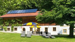 A building with a red-tiled roof, a white facade, and a large tree in front. There are outdoor seating areas with chairs and a yellow umbrella. - "Zellerhorn" Ferienwohnung I°, 39 qm, extra Schlafzimmer° (Aschau im Chiemgau)