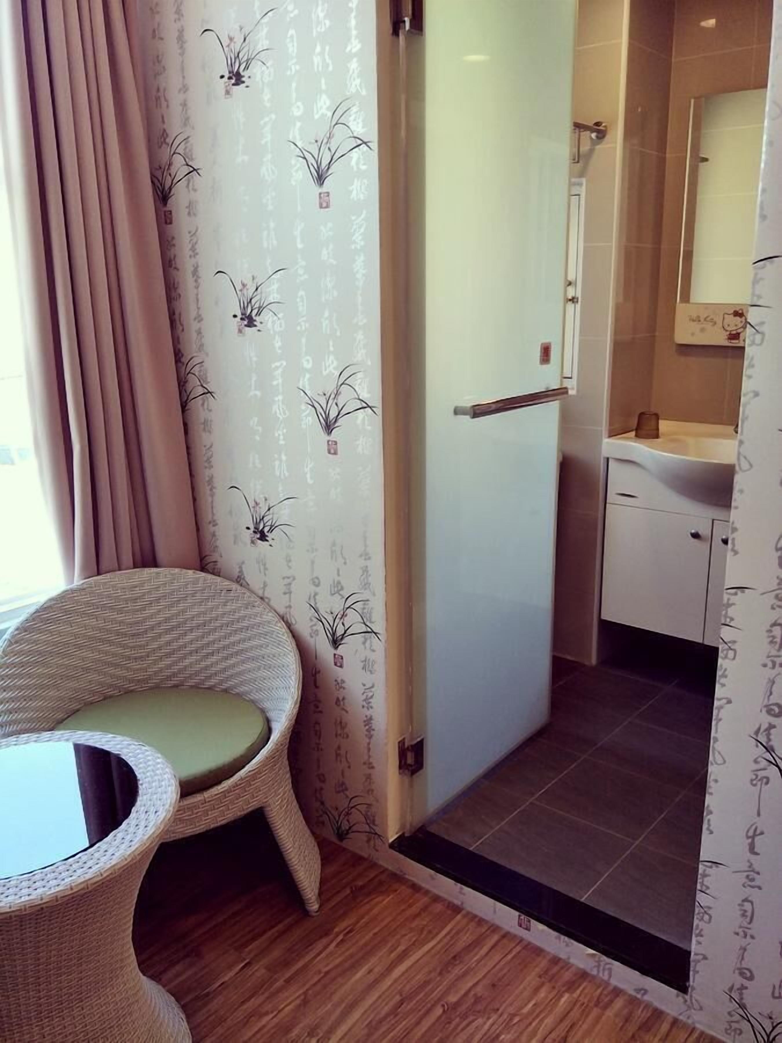 A room with a wicker chair, a small table, a shower area with a glass door, and a bathroom with a sink and mirror.