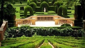 Un giardino formale con un vialetto centrale che conduce a una statua bianca, circondato da siepi curate e topiari.