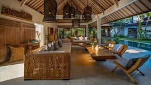 A spacious outdoor lounge with wicker furniture, hanging light fixtures, and a view of a swimming pool. - Abaca Villas (Seminyak)