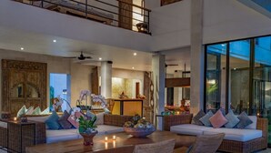 A spacious living area with a high ceiling, wicker furniture, and a large window providing a view of the outdoors. - Abaca Villas (Seminyak)