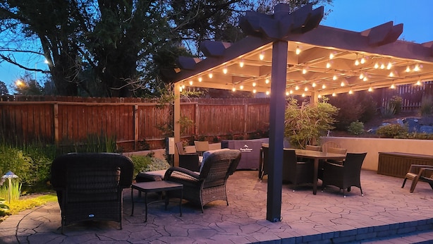 Un patio al aire libre con mobiliario de mimbre, una pérgola con luces colgantes y un suelo de baldosas de piedra.